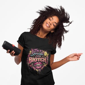 May include: Black t-shirt with a colorful graphic that reads "I'm a Sarcastic Little BIOTCH". The design includes a smiling face, lightning bolts, and a mouth with fangs. A black speaker is held in the left hand.