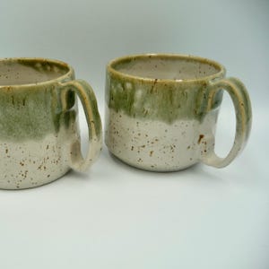 Peut inclure: Deux mugs en céramique faits à la main. Les mugs sont de couleur blanc cassé moucheté avec un glaçage vert qui s'estompe sur la moitié supérieure. Chaque mug a une grande anse incurvée.