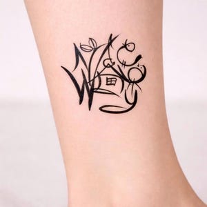 Op de afbeelding: Zwarte inkt tattoo op een enkel. De tattoo heeft een gestileerd ontwerp met de letter "W", een huis en bloemelementen. Het kunstwerk is een vrije hand stijl met een grillige esthetiek.