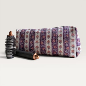 May include: A quilted fabric storage pouch featuring a repeating floral pattern in shades of purple, orange, and white, positioned next to a cylindrical hair styling tool with a metallic copper-colored top and a black perforated barrel.