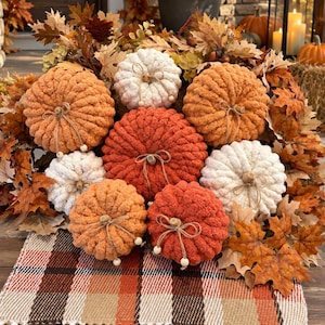 May include: A collection of seven plush, chunky knit pumpkins in shades of orange, rust, and cream, each topped with a twine bow and small wooden beads. The pumpkins are arranged on a plaid fabric runner with fringe, surrounded by autumn leaves.