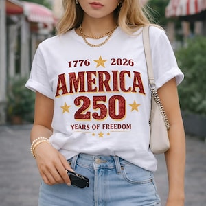 May include: White t-shirt with the text "1776 2026 AMERICA 250 YEARS OF FREEDOM" in red and gold. The shirt has a crew neck and short sleeves. The model is wearing blue jeans and holding sunglasses.