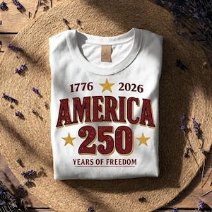 May include: White t-shirt with the text "1776 2026 AMERICA 250 YEARS OF FREEDOM" in burgundy and gold. The design includes gold stars. The shirt is folded on a woven mat.