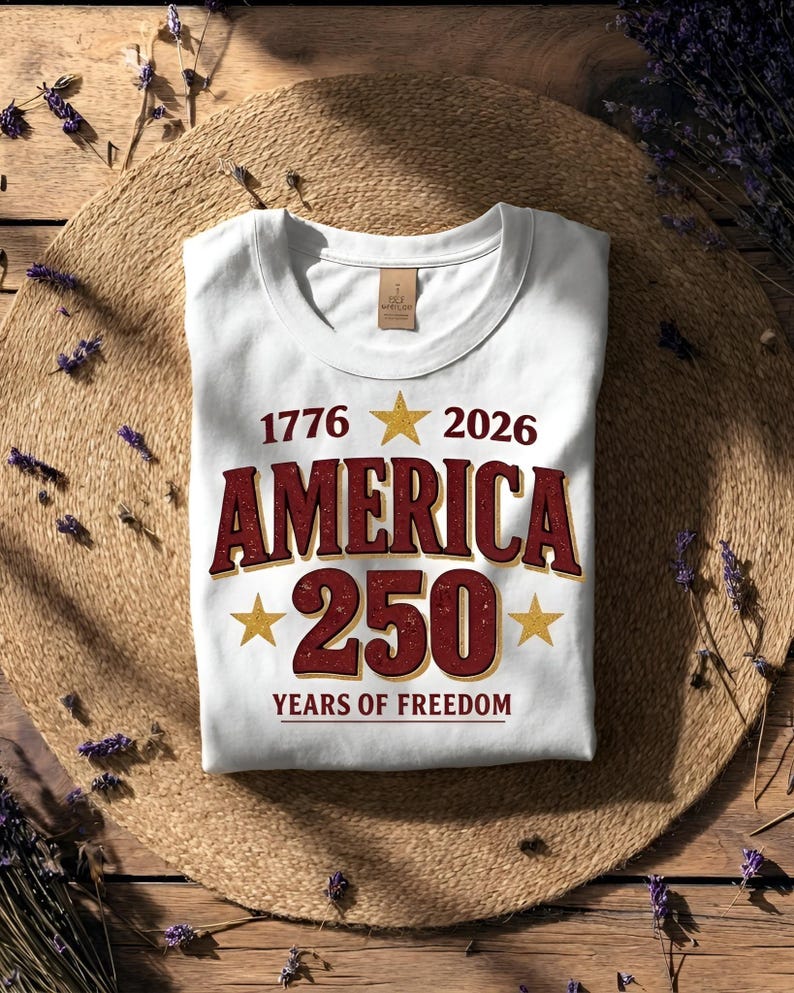 May include: White t-shirt with the text "1776 2026 AMERICA 250 YEARS OF FREEDOM" in burgundy and gold. The design includes gold stars. The shirt is folded on a woven mat.