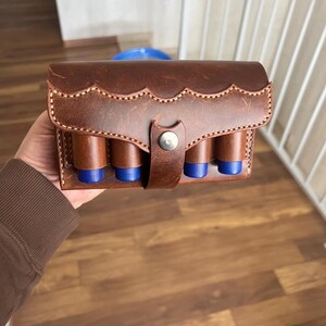 Puede incluir: Un portacartuchos de cuero marrón con borde festoneado y cierre de botón plateado. El soporte contiene cuatro tubos marrones con tapas azules, probablemente para munición. La costura es visible.