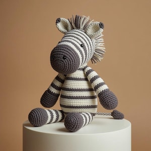 Può includere: Un peluche a forma di zebra realizzato all'uncinetto con un motivo a righe grigio scuro e crema. Il giocattolo presenta un muso arrotondato, due orecchie dritte, una corta criniera di frange di filato e una lunga coda con un ciuffo finale.