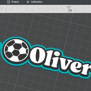 Könnte beinhalten: Ein weißes und schwarzes Fußball-Design mit dem Namen "Oliver" in fetter, weißer Schrift. Das Design ist türkis umrandet und hat oben eine kleine Schlaufe, was auf einen Schlüsselanhänger oder Anhänger hindeutet.