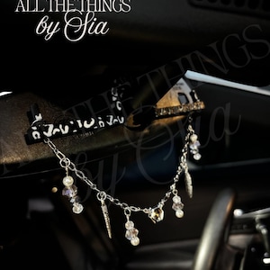 May include: A car rearview mirror charm featuring a black and white patterned bar with a silver-toned chain dangling below. The chain is adorned with various beads, including white pearls and clear crystals, along with small silver-toned metal charms.