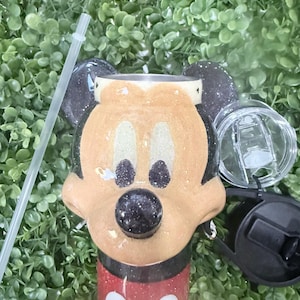 Op de afbeelding: Een tumbler met een Mickey Mouse-ontwerp. De tumbler heeft een rode basis met witte stippen en een glinsterend gouden gezicht. Zwarte oren en een zwarte neus maken het iconische karakter compleet. Een doorzichtig rietje en deksel zijn inbegrepen.
