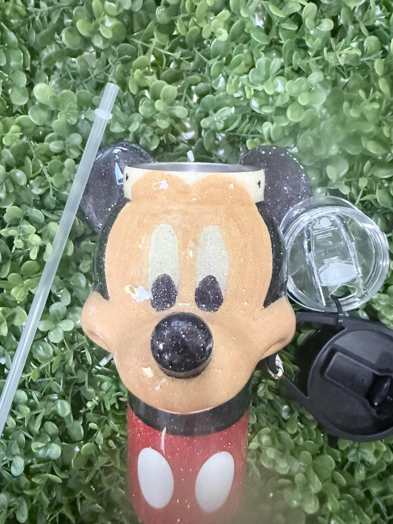 Op de afbeelding: Een tumbler met een Mickey Mouse-ontwerp. De tumbler heeft een rode basis met witte stippen en een glinsterend gouden gezicht. Zwarte oren en een zwarte neus maken het iconische karakter compleet. Een doorzichtig rietje en deksel zijn inbegrepen.