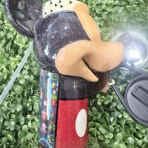 Op de afbeelding: Een zwart en rode glitter beker in de vorm van Mickey Mouse. De beker heeft een zwart deksel en een rode basis met een witte cirkel. De beker heeft een rietje en een zwarte zonnebril.