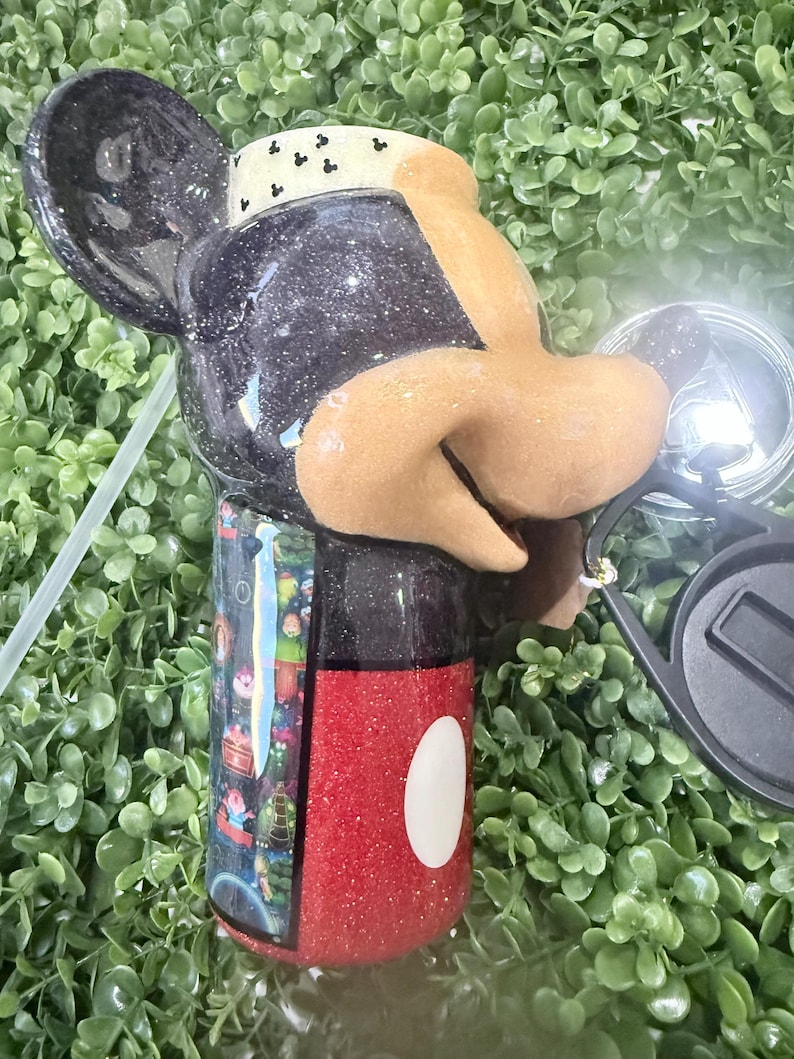 Op de afbeelding: Een zwart en rode glitter beker in de vorm van Mickey Mouse. De beker heeft een zwart deksel en een rode basis met een witte cirkel. De beker heeft een rietje en een zwarte zonnebril.
