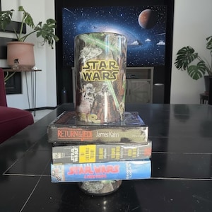 May include: A Star Wars themed glass tumbler sits atop a stack of VHS tapes. The tumbler features the Star Wars logo and images of Darth Vader and Stormtroopers. The VHS tapes include "Return of the Jedi" and "Star Wars" titles.