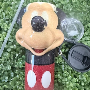 Op de afbeelding: Een glitterende beker met een Mickey Mouse-ontwerp. De beker heeft een rode basis met witte cirkels, een zwarte bovenkant en een bruin gezicht met een zwarte neus. Inclusief een doorzichtig deksel, zwarte handgreep en een doorzichtig rietje.