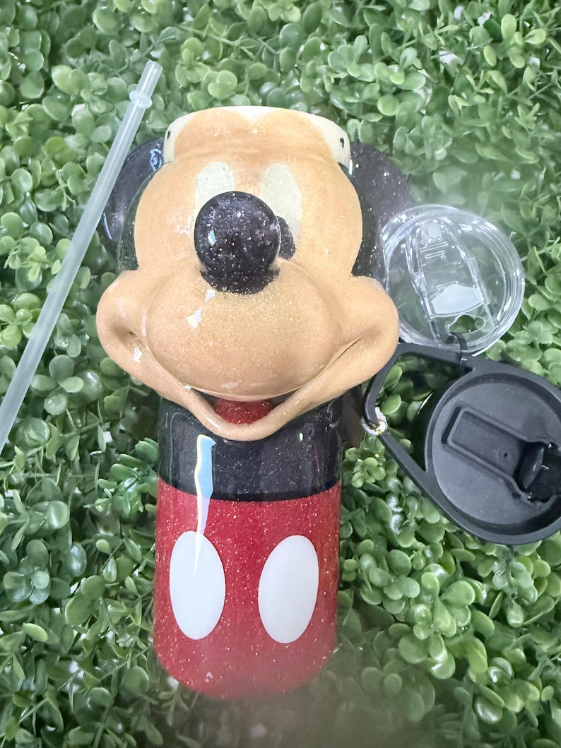 Op de afbeelding: Een glitterende beker met een Mickey Mouse-ontwerp. De beker heeft een rode basis met witte cirkels, een zwarte bovenkant en een bruin gezicht met een zwarte neus. Inclusief een doorzichtig deksel, zwarte handgreep en een doorzichtig rietje.