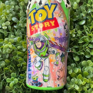 Peut inclure: Un gobelet avec des personnages du film d'animation "Toy Story". Le design comprend Buzz l'&Eacute;clair, les mots "Toy Story" et d'autres personnages. Le haut du gobelet pr&eacute;sente un motif tachet&eacute; blanc et noir.