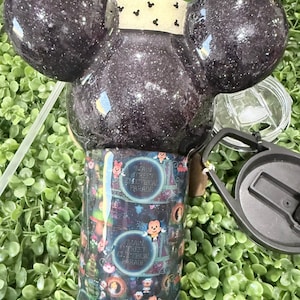 Op de afbeelding: Een glinsterende zwarte beker met Mickey Mouse oren. De beker heeft een kleurrijke wikkel met de tekst "Main Street Electrical Parade" en karakterillustraties. Inclusief deksel en rietje.
