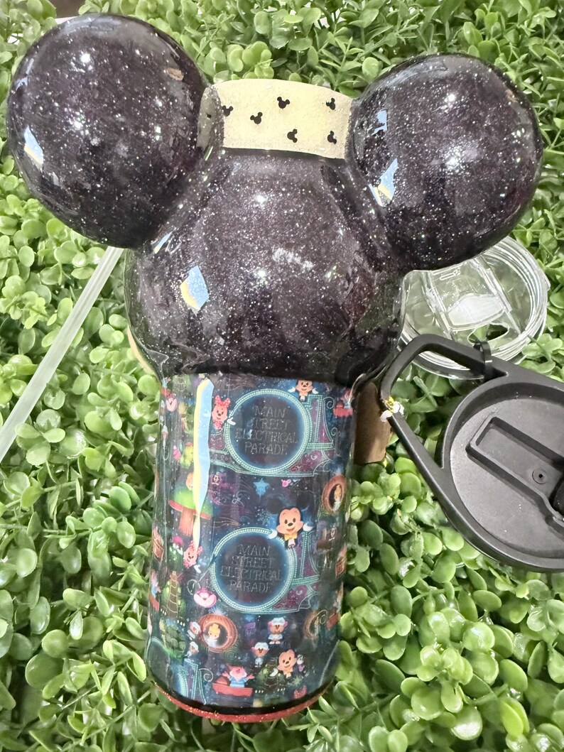 Op de afbeelding: Een glinsterende zwarte beker met Mickey Mouse oren. De beker heeft een kleurrijke wikkel met de tekst "Main Street Electrical Parade" en karakterillustraties. Inclusief deksel en rietje.