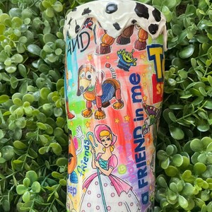 Peut inclure: Un gobelet color&eacute; avec des personnages du film d'animation "Toy Story". Le design comprend des images de Woody, Buzz l'&Eacute;clair, Bo Peep et d'autres personnages, avec les mots "A Friend in Me" et "Andy" visibles. Le haut du gobelet a un motif tachet&eacute; blanc et noir.