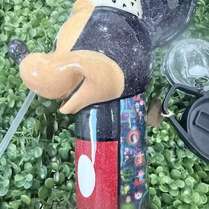 Op de afbeelding: Een glinsterende zwarte en rode beker in de vorm van Mickey Mouse. De beker heeft een wit rietje en een doorzichtig deksel. Het ontwerp omvat een witte en zwarte band rond het hoofd en een kleurrijke wikkel.