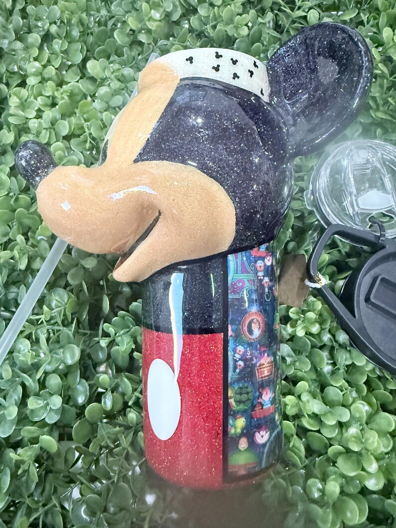 Op de afbeelding: Een glinsterende zwarte en rode beker in de vorm van Mickey Mouse. De beker heeft een wit rietje en een doorzichtig deksel. Het ontwerp omvat een witte en zwarte band rond het hoofd en een kleurrijke wikkel.