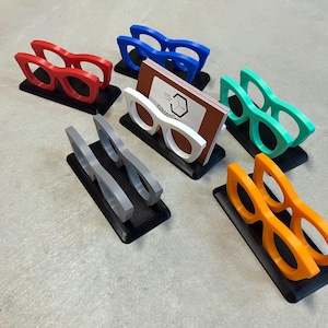 Glasses Business Card Holders sitting on concrete.  Colors are grey, blue, teal, red, white and orange.