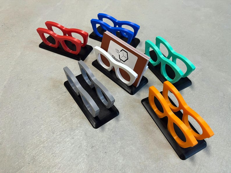 Glasses Business Card Holders sitting on concrete.  Colors are grey, blue, teal, red, white and orange.