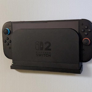 May include: A black Nintendo Switch console and dock are mounted on a white wall. The dock features the Nintendo Switch logo and the number 2. The controllers have blue and red accents.