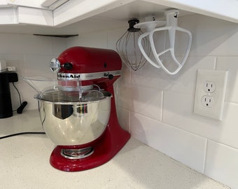 Op de afbeelding: Een rode KitchenAid-standmixer met een zilveren kom en een doorzichtige kunststof spatbeschermer. De mixer staat op een wit aanrecht met een witte tegelachterwand. Er hangen witte hulpstukken aan het kastje boven de mixer.