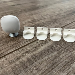 May include: Five white wall mounts for a smart home device, with one white smart home device on a stand in the background.