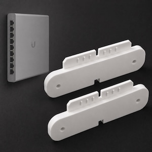 Könnte beinhalten: Ein hellgrauer Netzwerk-Switch mit mehreren Anschlüssen ist auf einer dunklen Oberfläche montiert. Zwei weiße Montagehalterungen sind darunter positioniert. Die Halterungen haben ein einzigartiges Design mit mehreren Schlitzen und Löchern, wahrscheinlich zur Sicherung des Switches.