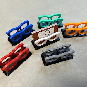 Glasses Business Card Holders sitting on concrete.  Colors are grey, blue, teal, red, white and orange.