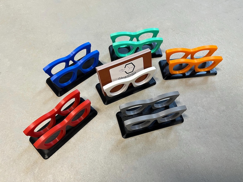Glasses Business Card Holders sitting on concrete.  Colors are grey, blue, teal, red, white and orange.