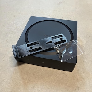 Op de afbeelding: Een zwarte Sonos-luidspreker met een wandbeugel en schroeven in een plastic zak. De luidspreker heeft een cirkelvormig ontwerp met een klein sleutelgat in het midden.