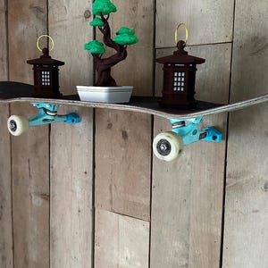 May include: A skateboard shelf with a black top, light blue trucks, and white wheels. It holds a small bonsai tree in a white pot and two brown decorative lanterns with gold handles. The shelf is mounted on a wooden wall.