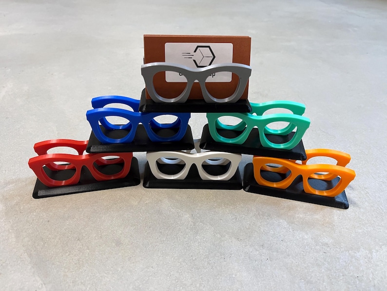 Glasses Business Card Holder stacked in a triangle sitting on concrete.  Colors are grey, blue, teal, red, white and orange.