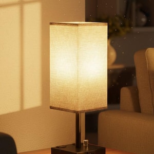 May include: A rectangular table lamp with a beige fabric shade. The lamp has a dark metal base with a power outlet and a USB port. The lamp is on a wooden table in a room with warm lighting.
