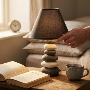 May include: A stone-stacked lamp with a brown lampshade is on a wooden table beside an open book and a coffee mug. The lamp base is made of various sized stones. A hand touches the lampshade. A clock is in the background.