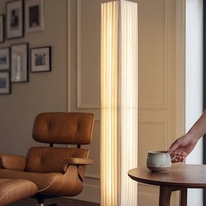Peut inclure: Un lampadaire rectangulaire et élancé avec un abat-jour texturé de couleur claire et une base en bois. La lampe est allumée, diffusant une lueur chaleureuse. Un fauteuil en cuir marron et une petite table en bois avec une tasse sont également visibles.
