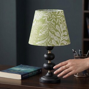 Peut inclure: Une petite lampe de table avec un abat-jour à motifs floraux verts et blancs. La lampe a une base noire et un corps en bois empilé avec des accents en laiton. Un livre et un porte-stylo sont également visibles.