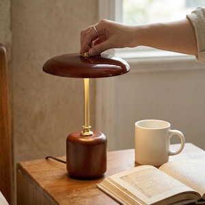Op de afbeelding: Een houten tafellamp met een donkerbruine lampenkap en voet, en een goudkleurige steel. Een witte mok en een open boek liggen op de houten tafel. De lamp wordt door een hand aangezet.