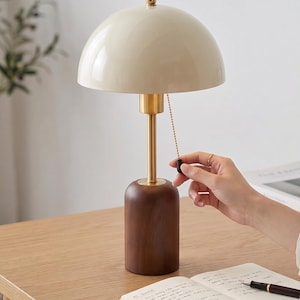 May include: A modern desk lamp with a cream-colored dome shade, brass stem, and a dark wood base. The lamp has a pull chain and sits on a wooden desk next to a notebook and pen. The lamp is a stylish addition to any home office.