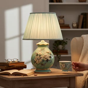Peut inclure: Une lampe de table décorative avec un abat-jour vert clair et un pied en céramique. Le pied est de couleur vert clair avec un motif floral. La lampe est sur une table en bois avec des livres et une tasse à thé.