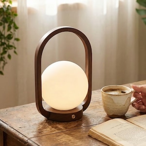 Puede incluir: Una lámpara de mesa de madera con una pantalla esférica blanca brillante. La lámpara tiene un marco de madera arqueado de color marrón oscuro. Una taza de café y un libro abierto están sobre la mesa de madera. Un artículo de decoración.