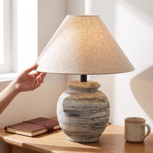 Peut inclure: Une lampe de table avec un abat-jour en lin beige et un pied en céramique avec un motif tourbillonnant gris et blanc. La lampe est posée sur une table en bois, avec un journal en cuir marron et une tasse beige.