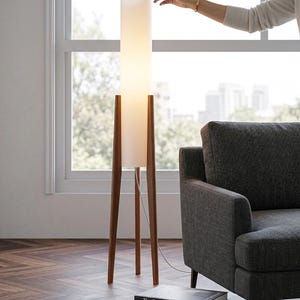 Op de afbeelding: Een moderne vloerlamp met een hoge, cilindrische witte kap en houten driepootpoten. De lamp staat bij een raam en een grijze bank. Een stapel boeken met de titels "The Power of Light" en "Architecture of Cities" is zichtbaar.