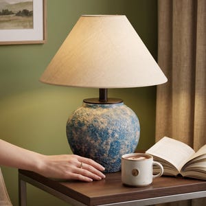 Peut inclure: Une lampe de table avec un abat-jour crème et une base en céramique texturée bleue et blanche. Une tasse de café et un livre ouvert sont posés sur une table marron foncé. La lampe est allumée.