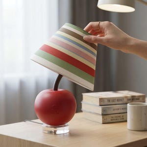 Peut inclure: Lampe de table originale en forme de pomme rouge avec un abat-jour rayé multicolore. La lampe a une base transparente et une tige marron foncé. Une main tient l'abat-jour.