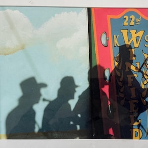 May include: A framed photograph featuring shadows of people and a colorful sign. The sign reads "22nd KWS FIELD" in yellow lettering against a red, blue, and green background. Shadows of figures in hats are cast on a light blue backdrop.