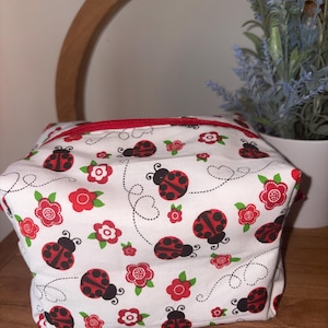 May include: A white fabric pouch featuring a repeating pattern of red and black ladybugs, small red flowers with white centers, green leaves, and thin black dashed lines. The pouch has a red zipper closure along the top edge.